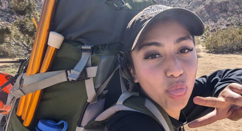 A person wearing a backpack makes a silly face and gives the camera a peace sign. 